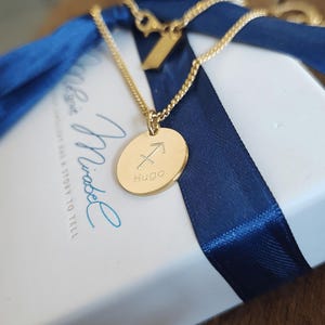 May include: A gold-colored necklace with a round pendant featuring the Sagittarius symbol and the name "Hugo". The necklace is on a white box with a blue ribbon. The brand name "Mirabel" is visible on the box.