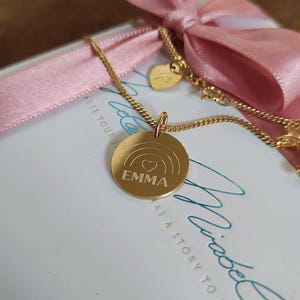 May include: A gold-colored necklace with a circular pendant featuring a rainbow design and the name "EMMA" engraved in the center. The necklace is set against a backdrop of a white box with a pink ribbon and a heart-shaped charm.