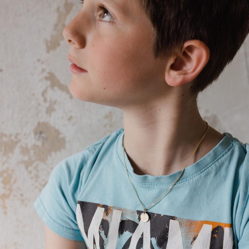 May include: A gold-colored necklace with a small, round pendant is worn over a light blue t-shirt. The t-shirt has a graphic print with the word "MIAMI" visible. The necklace hangs on the person's chest.