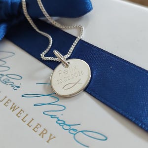May include: A silver pendant necklace with a round charm. The charm is engraved with the name "Felix", the date "22.02.2026", and a fish symbol. The necklace is on a blue ribbon and a white box.