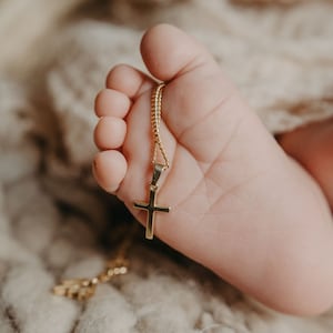 May include: A gold chain necklace with a small gold cross pendant resting on a baby's foot.