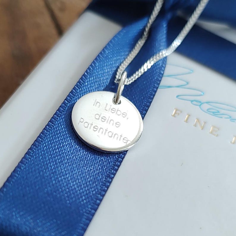 May include: A silver pendant necklace with a round charm. The charm has engraved text and hangs on a delicate chain. The necklace rests on a white surface with a blue ribbon. The text on the charm reads "In Liebe, deine Patentante."