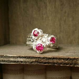 以下が含まれることがあります： 花の形をしたセッティングに3つのピンクの宝石がセットされたシルバーリング。リングはテクスチャ加工が施され、繊細なデザインです。