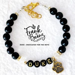 May include: Black beaded bracelet with a gold charm that says "DUKE" and a gold paw print charm. The bracelet has a gold clasp and clear beads.