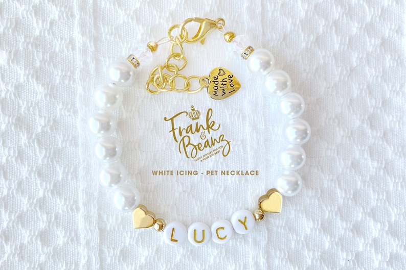 May include: White pearl necklace with a gold charm that says "Frank & Beans" and a heart charm that says "Made with Love". The necklace also has a gold heart charm and a white bead with the name "Lucy" spelled out in gold letters.