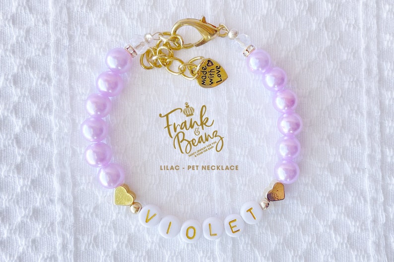 May include: Lilac pet necklace with gold heart charms and a name tag that reads "VIOLET". The necklace has a gold clasp and a charm that says "Made with Love".