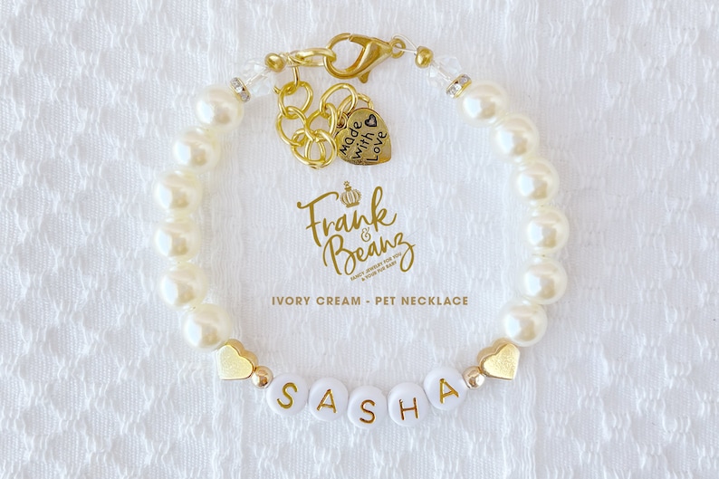 May include: Ivory cream pet necklace with gold heart charms and a name tag that reads "SASHA". The necklace also features a gold charm with the text "Made with love".