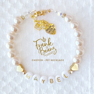 May include: White pearl pet necklace with a gold heart charm that says "Made with Love" and a gold charm with the text "Frank & Beanz" and a crown. The necklace also has gold heart beads and white beads with the name "MAYBEL" spelled out.