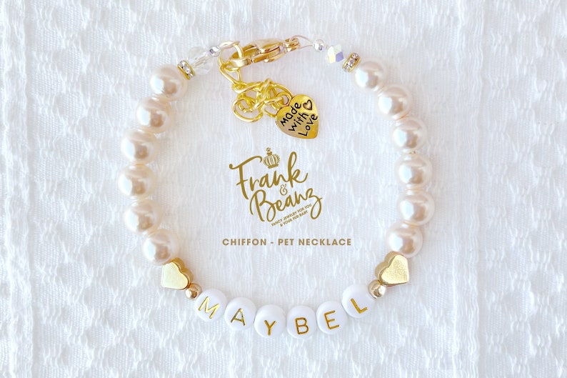 May include: White pearl pet necklace with a gold heart charm that says "Made with Love" and a gold charm with the text "Frank & Beanz" and a crown. The necklace also has gold heart beads and white beads with the name "MAYBEL" spelled out.