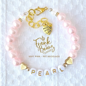 May include: Pink pearl pet necklace with a gold chain and a heart charm that says "Made with Love". The necklace also has a charm with the text "Frank & Beanz" and a charm with the name "PEARL".