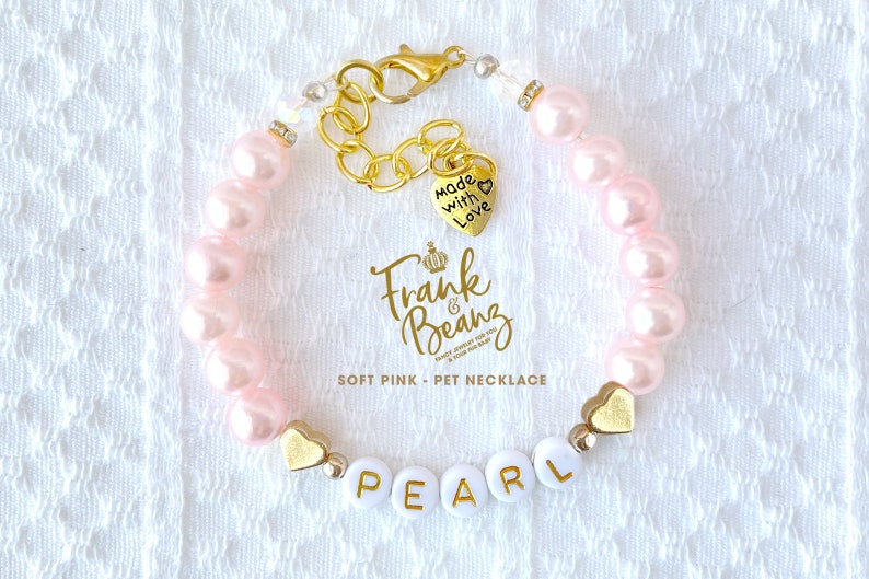 May include: Pink pearl pet necklace with a gold chain and a heart charm that says "Made with Love". The necklace also has a charm with the text "Frank & Beanz" and a charm with the name "PEARL".