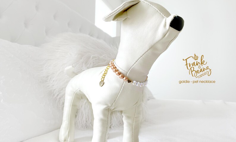 White dog mannequin wearing a champagne pearl dog necklace with 14K gold heart accents, personalized with gold and white letter beads spelling “GOLDIE”. Chain extender with a heart charm engraved with "made with love", standing on a white bed in a bright bedroom.