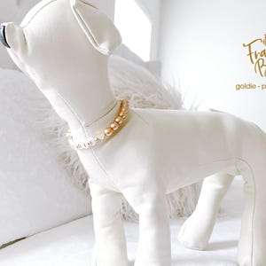 White dog mannequin wearing a champagne pearl dog necklace with 14K gold heart accents, personalized with gold and white letter beads spelling “GOLDIE,” standing on a white bed in a bright bedroom.