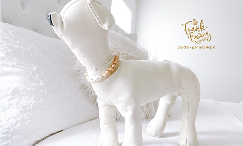 White dog mannequin wearing a champagne pearl dog necklace with 14K gold heart accents, personalized with gold and white letter beads spelling “GOLDIE,” standing on a white bed in a bright bedroom.
