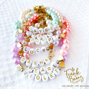 May include: A set of colourful pearl bracelets with gold heart charms and white letter beads spelling out names such as "Maybel", "Chase", "Poppy", "Lucy", "Sasha", "Pearl", and "Violet". The bracelets are arranged in a stack and feature a "Frank & Beanz" logo with the text "14K Heart - Pet Necklace".