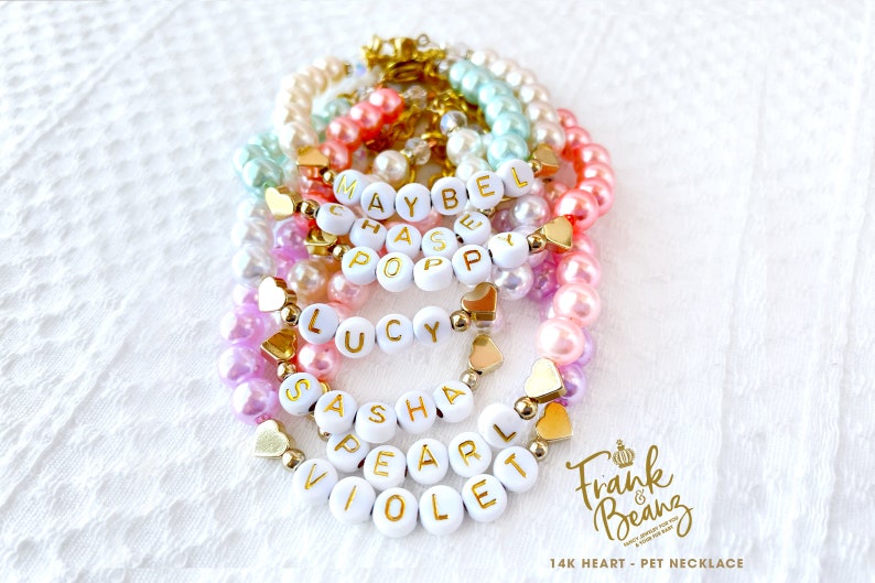 May include: A set of colourful pearl bracelets with gold heart charms and white letter beads spelling out names such as "Maybel", "Chase", "Poppy", "Lucy", "Sasha", "Pearl", and "Violet". The bracelets are arranged in a stack and feature a "Frank & Beanz" logo with the text "14K Heart - Pet Necklace".