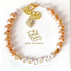 Champagne pearl pet necklace with 14K gold heart accents and a dangling “made with love” charm, personalized with white and gold letter beads spelling “GOLDIE,” displayed on a white gift box.