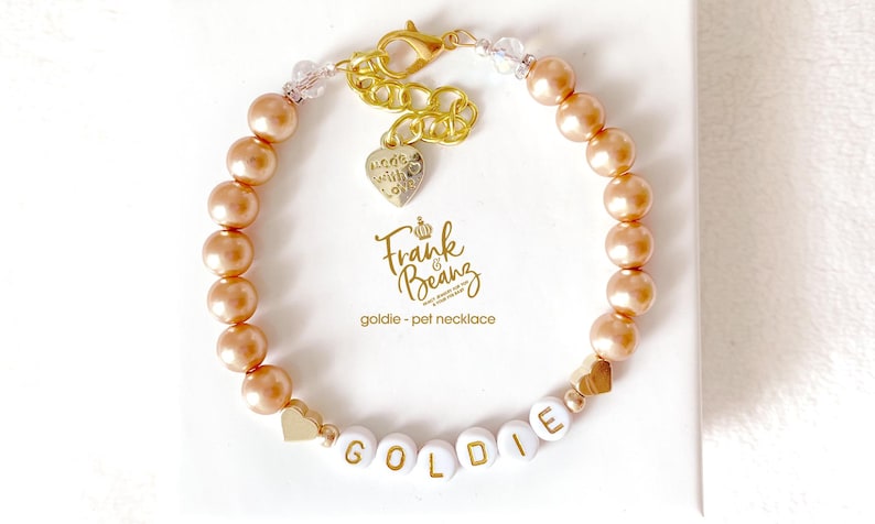 Champagne pearl pet necklace with 14K gold heart accents and a dangling “made with love” charm, personalized with white and gold letter beads spelling “GOLDIE,” displayed on a white gift box.