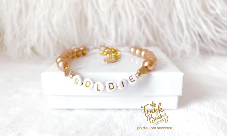 Champagne pearl pet necklace with 14k gold heart charms, personalized with white and gold letter beads that spell the name 'GOLDIE'. The necklace is on a white gift box with Frank & Beanz logo.
