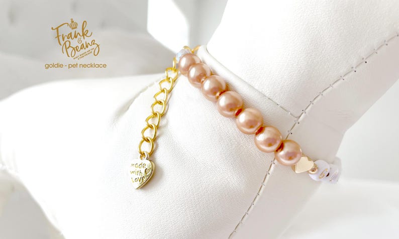 Up close white dog mannequin wearing a champagne pearl dog necklace with 14K gold heart accents with a dangling gold chain attached to a gold heart charm with the engraved words "made with love".