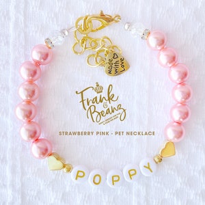 May include: A pink pearl pet necklace with a gold heart charm and the name "Poppy" spelled out in white letters with gold lettering. The necklace has a gold clasp and a small gold charm that says "Made with Love".
