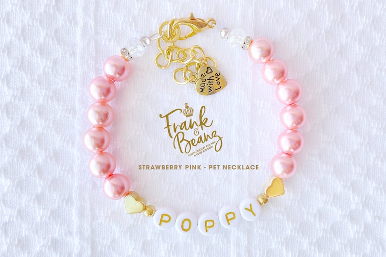 May include: A pink pearl pet necklace with a gold heart charm and the name "Poppy" spelled out in white letters with gold lettering. The necklace has a gold clasp and a small gold charm that says "Made with Love".