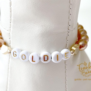 Close-up of a champagne pearl dog necklace with 14K gold heart accents and white letter beads spelling “GOLDIE,” shown on a white dog mannequin.