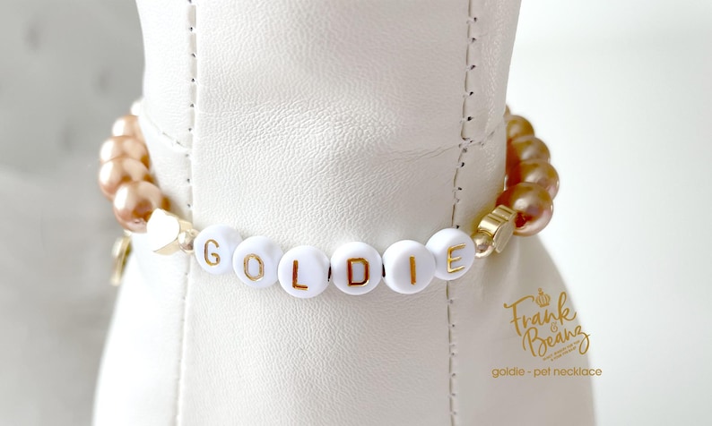 Close-up of a champagne pearl dog necklace with 14K gold heart accents and white letter beads spelling “GOLDIE,” shown on a white dog mannequin.