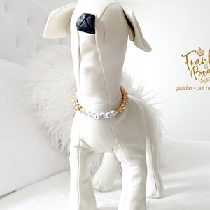 White stuffed dog mannequin wearing a champagne pearl dog necklace with 14K gold heart accents, personalized with white and gold letter beads spelling “GOLDIE,” posed on a white bed background.