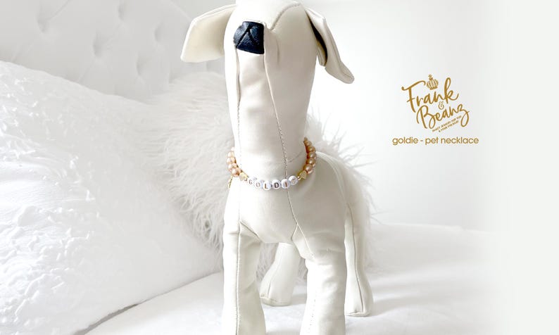White stuffed dog mannequin wearing a champagne pearl dog necklace with 14K gold heart accents, personalized with white and gold letter beads spelling “GOLDIE,” posed on a white bed background.