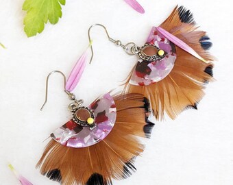 May include: A pair of earrings with brown feathers and a pink and white patterned resin piece. The resin piece has a small silver charm with a red stone.