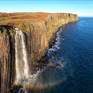 May include: A waterfall plunges down a dramatic cliff face, forming a rainbow over the sea. The cliff is rugged and brown, with green vegetation at the top. The sea is a deep blue, with whitecaps on the waves.