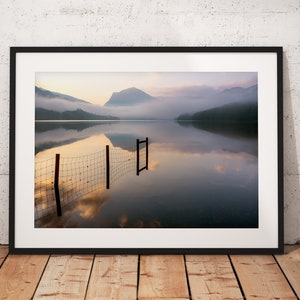 May include: A photograph of a lake with a mountain in the background. The lake is calm and the water is reflecting the sky and the mountain. There is a fence in the water, and the fence is reflected in the water as well.