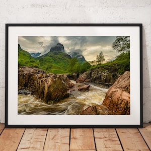 May include: A scenic landscape photograph featuring a rushing river flowing through a rocky gorge. The river is surrounded by lush green vegetation and towering mountains in the background. The sky is overcast with clouds, creating a dramatic and moody atmosphere.