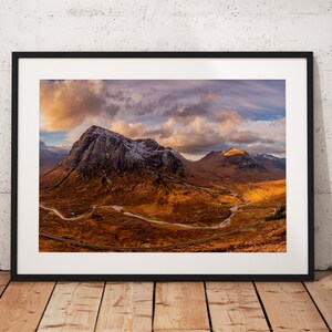 May include: A panoramic view of a mountain range with a winding road leading through the landscape. The mountains are covered in snow and the sky is a mix of blue and grey with clouds.