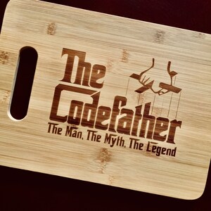 May include: A rectangular bamboo cutting board with a handle cutout. The board features the engraved text "The Codefather: The Man, The Myth, The Legend" along with a graphic of a hand holding puppet strings. The board is light brown.