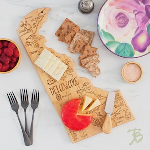 May include: A wooden serving board in the shape of Delaware, with etched city names and a map design. The board holds cheese, crackers, and a red cheese wheel. Includes a cheese knife, forks, and a bowl of raspberries.