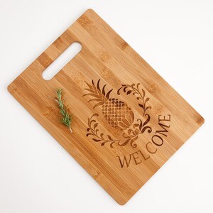 Pu&ograve; includere: Un tagliere in legno con un design a ananas e la parola "WELCOME" incisa su di esso. Il tagliere ha un manico ed &egrave; fatto di bamb&ugrave;.