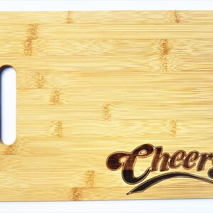 May include: A rectangular bamboo cutting board with a handle cutout on the left side. The word "Cheers" is etched in a script font on the lower right corner. The board has a light brown color.