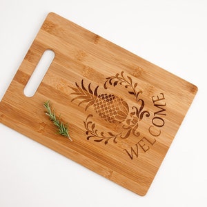 Pu&ograve; includere: Un tagliere in bamb&ugrave; naturale con un design a forma di ananas e la parola "WELCOME" incisa sulla superficie. Il tagliere ha un'apertura per la maniglia e un rametto di rosmarino &egrave; appoggiato sulla superficie.