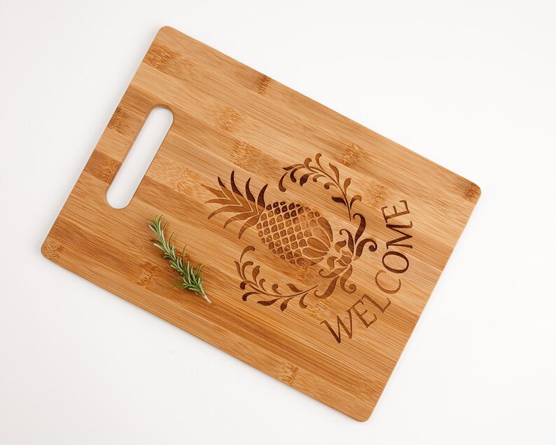 Pu&ograve; includere: Un tagliere in bamb&ugrave; naturale con un design a forma di ananas e la parola "WELCOME" incisa sulla superficie. Il tagliere ha un'apertura per la maniglia e un rametto di rosmarino &egrave; appoggiato sulla superficie.