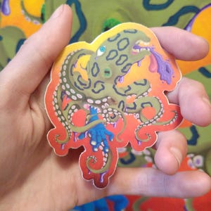 May include: A sticker of a green octopus with a purple tongue and blue eyes. The octopus is on a red, orange, and yellow background.