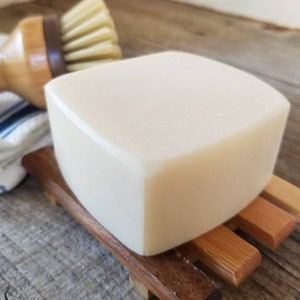 May include: A large, off-white bar of soap sits on a small, wooden soap dish. A wooden handled brush with light-colored bristles is in the background. A white and blue striped cloth is partially visible.