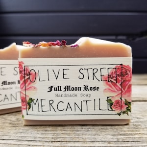 May include: A bar of handmade soap with a label that reads "Olive Street Mercantile Full Moon Rose." The soap is light pink with dried flower petals on top. The label features pink roses.