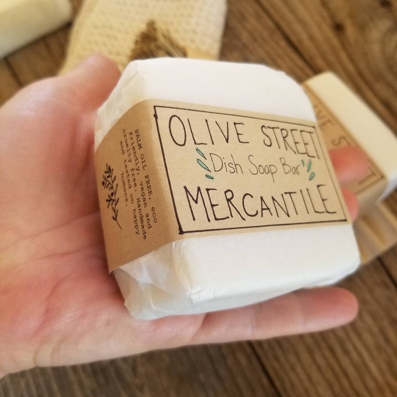 May include: A white dish soap bar wrapped in paper, held in a hand. The label reads "OLIVE STREET Dish Soap Bar MERCANTILE". The label also states the soap is palm oil free, vegan, and cruelty-free.
