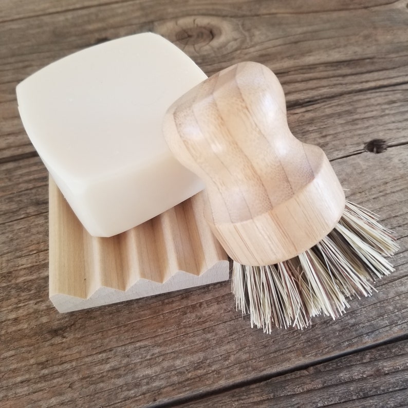 May include: A natural soap bar, wooden soap dish, and a wooden handled brush with bristles. The soap is a light color and sits on a grooved wooden dish. The brush has a rounded handle and natural bristles. The items are on a wooden surface.