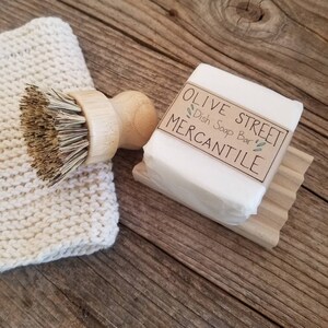 May include: A wooden handled dish brush with natural bristles, a white knitted washcloth, and a bar of dish soap on a wooden soap dish. The soap is labeled "Olive Street Dish Soap Bar Mercantile". The items are arranged on a rustic wooden surface.