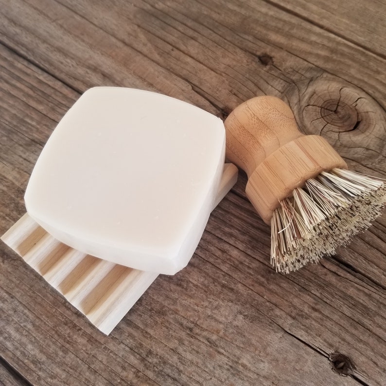 May include: A square, off-white bar of soap rests on a light wood soap dish. A wooden dish brush with natural bristles sits to the right. The items are arranged on a rustic, weathered wooden surface.