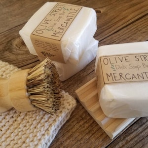 May include: White dish soap bars wrapped in paper, labeled "Olive Street Mercantile Dish Soap Bar." A wooden dish brush and a wooden soap dish are also present. The soap bars are on a wooden surface.