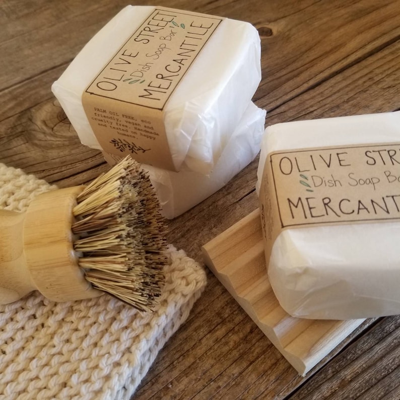 May include: White dish soap bars wrapped in paper, labeled "Olive Street Mercantile Dish Soap Bar." A wooden dish brush and a wooden soap dish are also present. The soap bars are on a wooden surface.